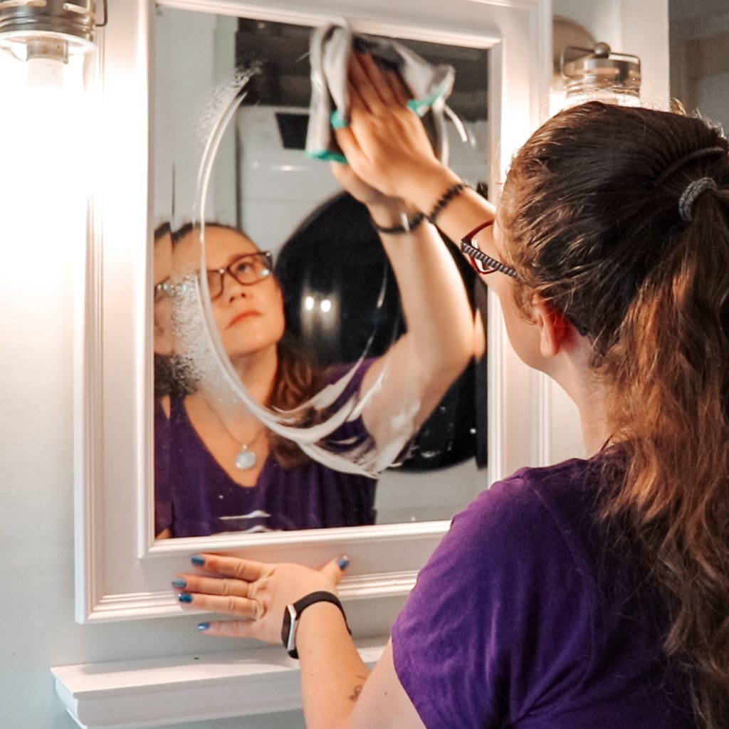 Jessica from The Clean Reset Co. cleaning a bathroom mirror, wearing a teal company T-shirt and gloves while wiping the glass with a microfiber cloth.