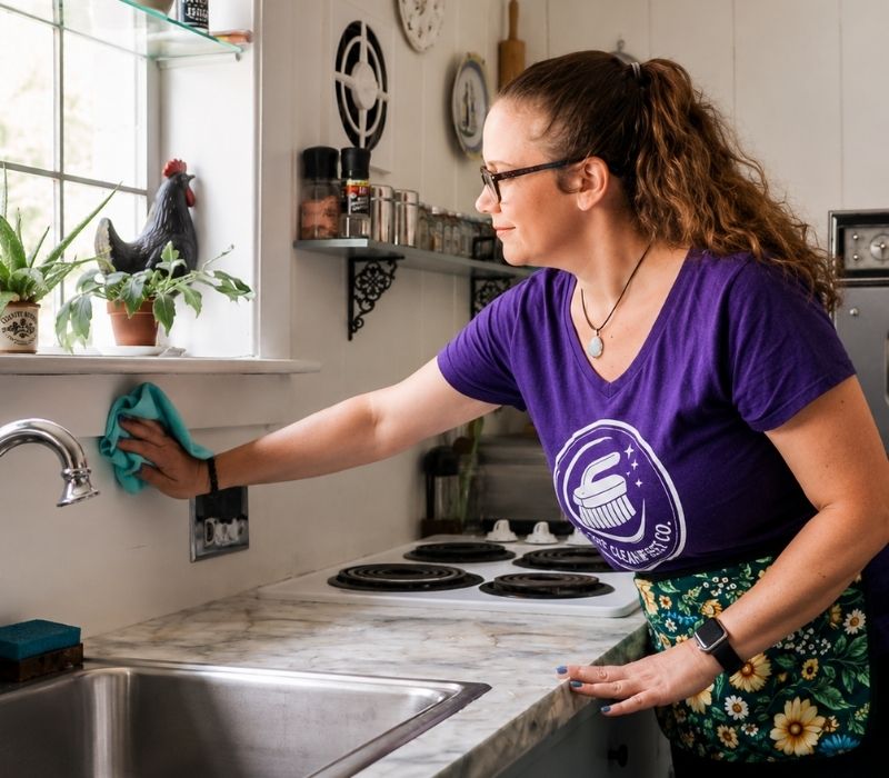 Solo cleaner Jessica from The Clean Reset Co. cleaning a kitchen counter, wearing a purple company T-shirt and gloves in a bright, tidy home.