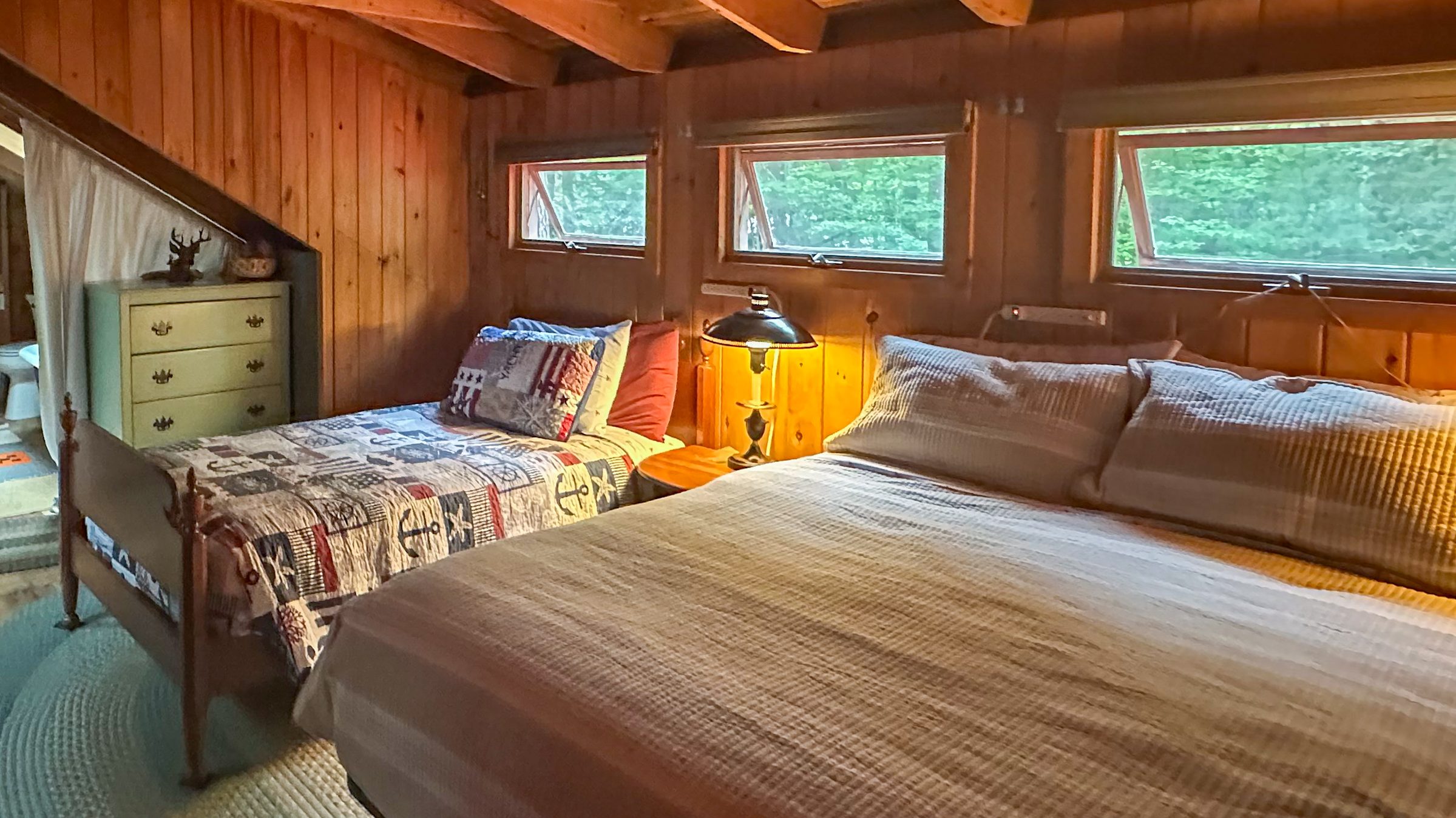 Cozy Maine camp bedroom with exposed wood beams, two neatly made beds, braided rugs, and wooded views through the windows.